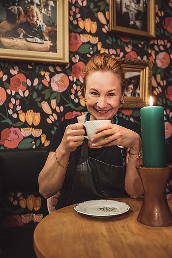 Madlén Sjödaardh med en kopp kaffe inne på Madrix. Framför syns ett stort, grönt ljus och i bakgrunden är tapeten svart och blommig. Det hänger foton i guldramar på väggen.
