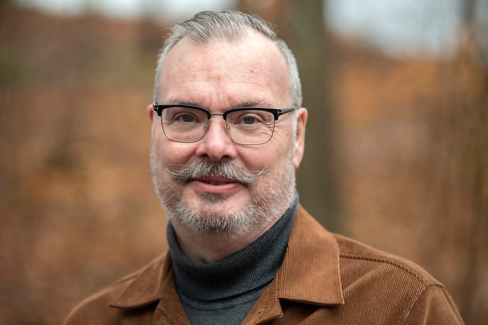 Porträttbild på Ulf Bengtsson - man med brun mustasch och glasögon klädd i brun kavaj, som ler och tittar in i kameran.
