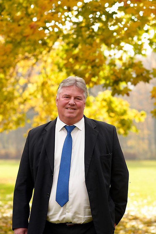 Bengt-Arne Persson i svart kostym, vit skjorta och blå slips mot höstlig bakgrund.